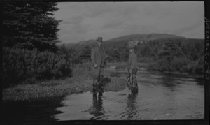 Image: Two men stand in water by trees, fishing